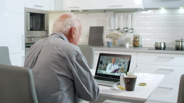 Medium shot of senior Caucasian man looking at laptop screen and telling about his health problems to online male doctor