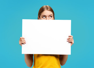 Funny girl with white empty poster, looking away, on blue background. Copy space for your text.