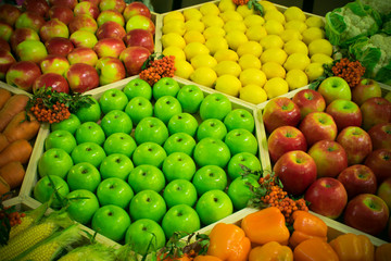 Photo of a counter on which green and red apples, lemons, bell pepper, carrots, corn and cauliflower are neatly laid out