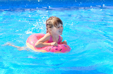 Fototapeta na wymiar Cute smiling little girl with inflatable donut circle learns to swim in swimming pool on hot sunny day. Healthy and happy childhood concept.