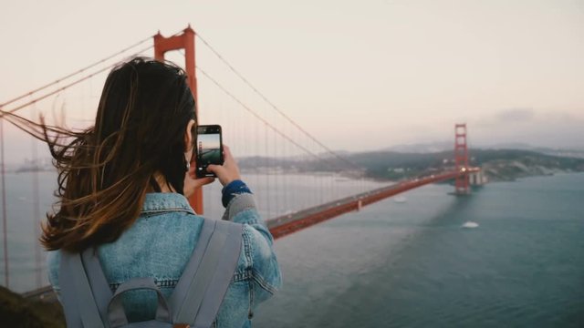 Back view young woman tourist with backpack, wind in hair takes smartphone photo of Golden Gate Bridge San Francisco.