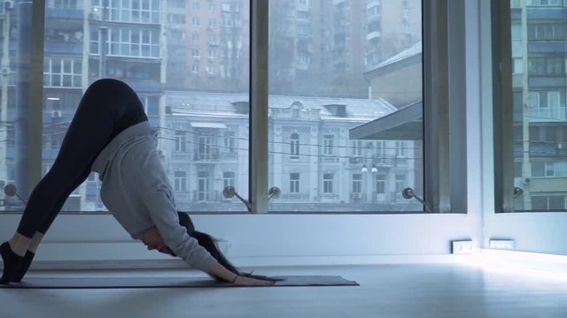 Young woman stretching legs on mat in the dancing hall with large floor-to-ceiling windows. Beautiful cityscape behind the glass. Girl exercising healthy lifestyle in fitness studio