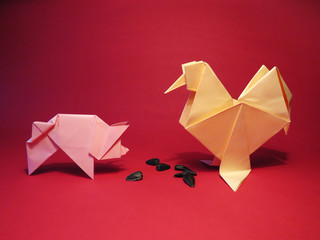 Yellow origami rooster with a pink pig and sunflower seeds on a red background