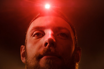 Close-up head-shot of young bearded extreme man with red head lamp on forehead