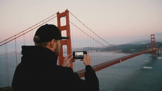 Back view young happy tourist man in black clothes takes phone photo of majestic sunset Golden Gate Bridge, California.