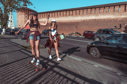 Two girls girlfriends summer city, fashionable students skateboard, longboard casual wear. Ride play rest in weekend fun day. They talk smiling street. Background road cars and sidewalk, free space.