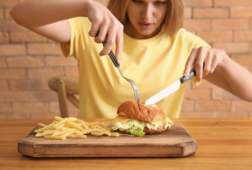 Young woman eating tasty burger with french fries at table