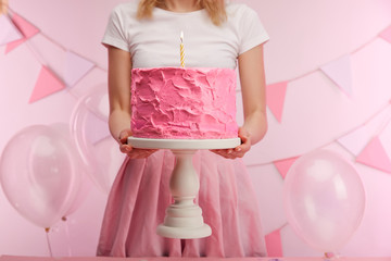 cropped view of woman holding cake stand with sweet pink birthday cake and burning candle near air balloons and decoration