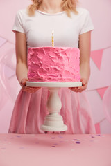 cropped view of woman holding cake stand with sweet pink birthday cake and burning candle