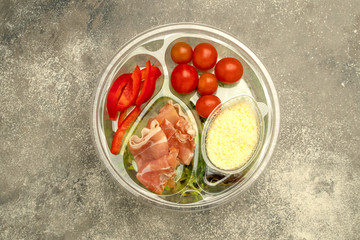 Healthy fast food in a transparent container. Vegetable salad for quick cooking with tomatoes, bell peppers, jamon and lettuce. On gray background. Top view.