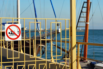 Fence on the pier with caution signs. Prohibition to swim and fish. Sailing yacht in the background.