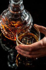 Whiskey glass hold by man hand and crystal decanter on dark background. Selective focus. Copy space.