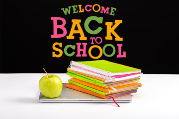 closed laptop, green apple, notebooks and color pencils on desk with welcome back to school lettering on black
