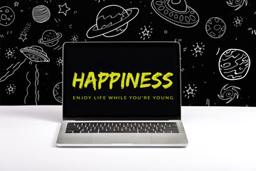 laptop on table with enjoy life while you are young and happiness lettering on screen with white galaxy illustration on black
