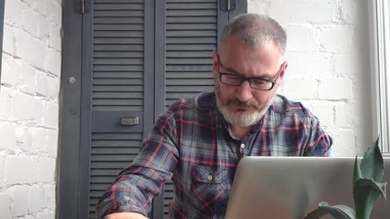 Gray-haired bearded male accountant working at home behind a laptop, making a report against the backdrop of a minimalist interior, checks data with the monitor, drinks tea or coffee and writes. - Powered by Adobe