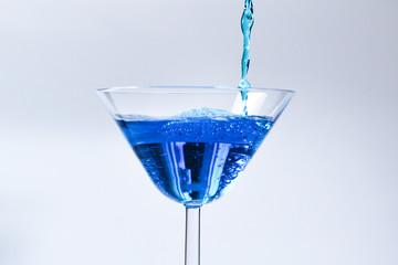 Cocktail with blue liquid in glass. Glass with blue water pouring with liquid with splashes and drops. Martini glass filling with alcohol with splashes on white background. Refreshing drink concept.