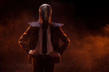 Kendo fighter in formal wear and helmet standing with arms akimbo on dark