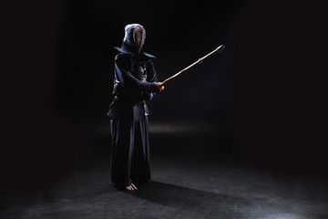 Full length view of kendo fighter in armor practicing with bamboo sword on black