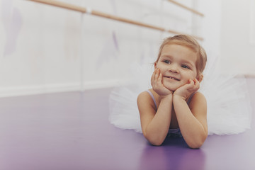 Cute happy litlle girl smiling looking away, cupping her face, copy space. Adorable little ballerina resting after ballet dance class at the studio. Carefree, childhood, happiness concept