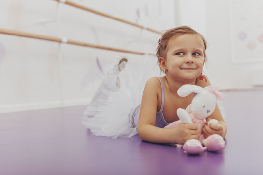 Happy little girl wearing tutu skirts and leotard lying on the floor with her plush rabbit toy, resting after ballet class. Adorable cheerful little ballerina relaxing at ballet dance studio
