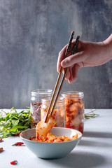 Korean traditional fermented appetizer kimchi cabbage and radish salad, fish snack served in glass jars with Vietnamese oregano and chili peppers over grey blue table. Chopsticks in mans hands. - obrazy, fototapety, plakaty