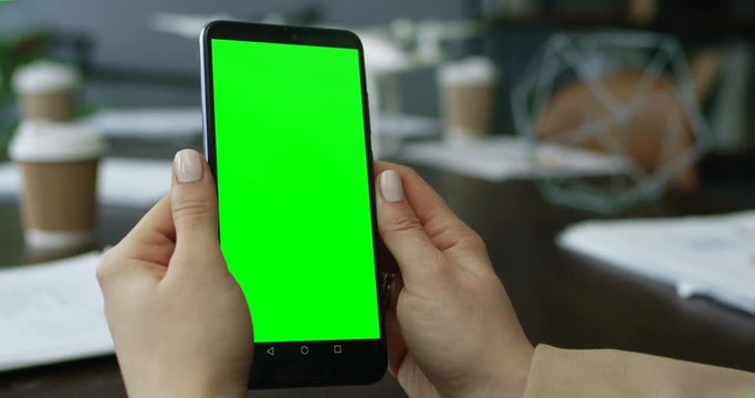 Close up of the Caucasian woman's hands holding black smartphone with chroma key screen in the office at the table during business meeting. Alpha channel.