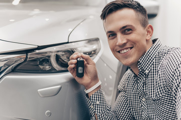 Close up of a happy young man smiling excitedly, holding car key, posing with his new automobile. Cheerful handsome male car owner at the dealership salon. Man buying new car. Ownership, rental 