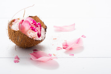 pile of pink bath salt with orchid flower petals at coconut shell bowl, white wood table