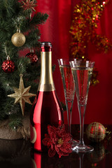 New Year. Red bottle and two champagne glasses are standing on a dark reflective surface near the New Year tree on a red background.