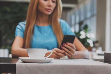 Selective focus on a coffee cup on the table on the foreground, beautiful long haired woman using her smart phone on the background. Woman messaging on her phone at the cafe. Technology, lifestyle