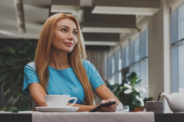 Gorgeous red haired woman using her smart phone at the restaurant while having her morning coffee. Cheerful beautiful woman smiling, looking away joyfully relaxing at the restaurant, copy space