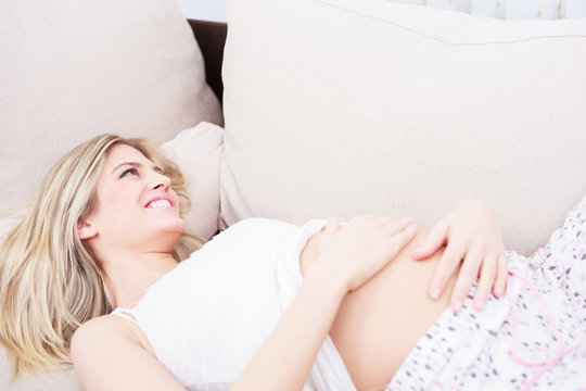 Happy pregnant young woman smiling and holding arms on her belly while lying on a sofa.