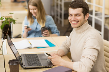 Handsome young cheerful man smiling to the camera using his smart phone while working on the laptop at the office copyspace casual creative designer businesspeople startup technology mobility carrier 