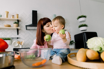 Funny little baby girl with mom on kitchen. Baby is sitting on the table and holding green apples while pretty young mother embracing and kissing her. Mother and daughter