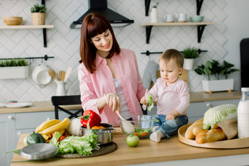 A young and beautiful mother in a pink shirt is preparing a fresh vegetable salad at home in the kitchen, together with her little cute daughter sitting on the table. mother and daughter