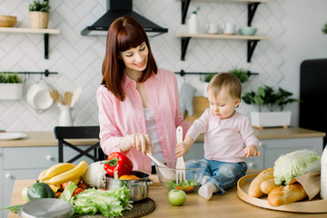 Cute little toddler baby girl holding wooden fork helping her pretty mother to mix salad at the table in the kitchen. Young beautiful woman with daughter cooking fresh salad at home.