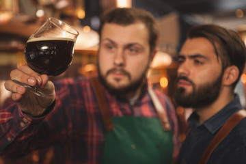 Selective focus on a glass of dark delicious beer in the hands of professional brewers. Two beermakers examining freshly brewed beer, working at their microbrewery, copy space. Business concept