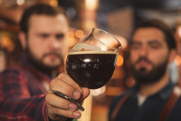 Close up shot of two brewers examining delicious dark beer in a glass, working at the beer production factory. Selective focus on a beer mug in the hands of beermakers. Quality, expertise concept - Powered by Adobe