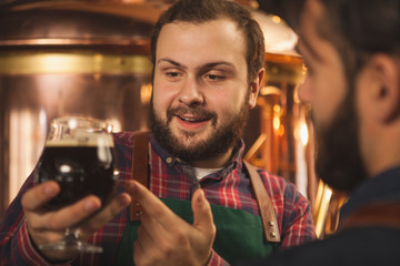 Close up shot of a cheerful bearded brewer smiling, talking to his colleague, examining dark beer in a glass. Two brewers producing craft beer at their microbrewery. Manufacturing, small business conc