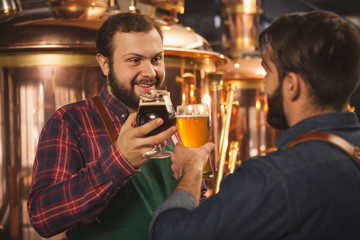 Bearded cheerful brewer talking to his assistant, working at the beer production production factory. Professional brewers clinking beer glasses, celebrating success. Small business, entrepreneurship c
