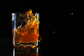 Ice cube falls into a glass with a whiskey on black background. Fluid in motion. Droplets of spray