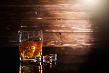 Glass of whiskey with ice cubes on black table against wooden background
