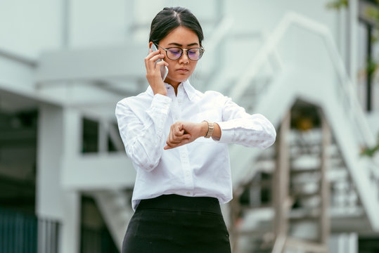 Asian businesswoman in eyeglasses talking business discussion with a smartphone while looking at her wristwatch for checking time standing front office building- woman officer worker and businesswoman