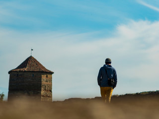 a man in a hooded sweater and knitted hat is walking along the road to the fortress against the background of the tower and the blue sky