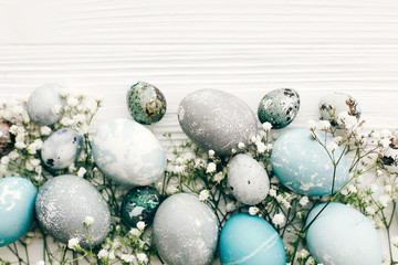 Fototapeta na wymiar Stylish Easter eggs with spring flowers border, flat lay on white wooden background with space for text. Modern easter eggs painted with natural dye in blue and grey marble. Happy Easter