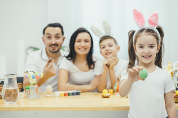 Pretty small girl with pigtails and bunny ears standing on the forefront, showing at the camera green easter egg. Her father mother and brother sitting at the table, preparing for Easter.