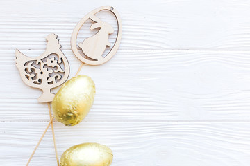 Stylish Easter chocolate eggs in golden foil and simple wooden bunny and chicken decorations on white wooden background, flat lay with space for text. Happy Easter. Holiday gift