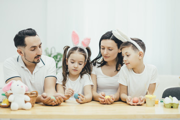 Friendly family sitting together at the table, holding colored easter eggs in their hands, looking how lovely girl with pigtails and bunny ears is colouring her blue easter egg.