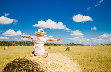 Naklejka na ściany i meble Hay balls on meadow on beautiful sunny day white clouds, adorable blond curly hear girl sit on the hay ball, hands reached out, happiness concept. Wearing hat, blue sky and forest in the distance.