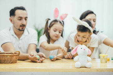 Fototapeta premium Mother father son and daughter involved in exciting colouring of easter eggs on the blurred background. Funny white rabbit on the forefront.